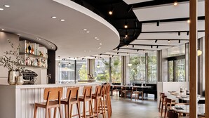 Uma área de bar moderna, com banquetas de madeira, um balcão branco e uma grande janela com vista para o exterior.