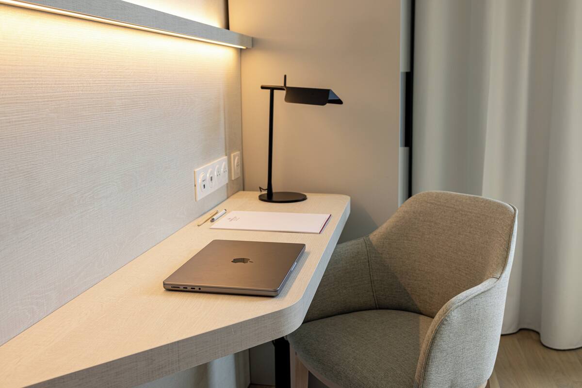 A modern hotel room with a desk, laptop, lamp, and a grey upholstered chair.