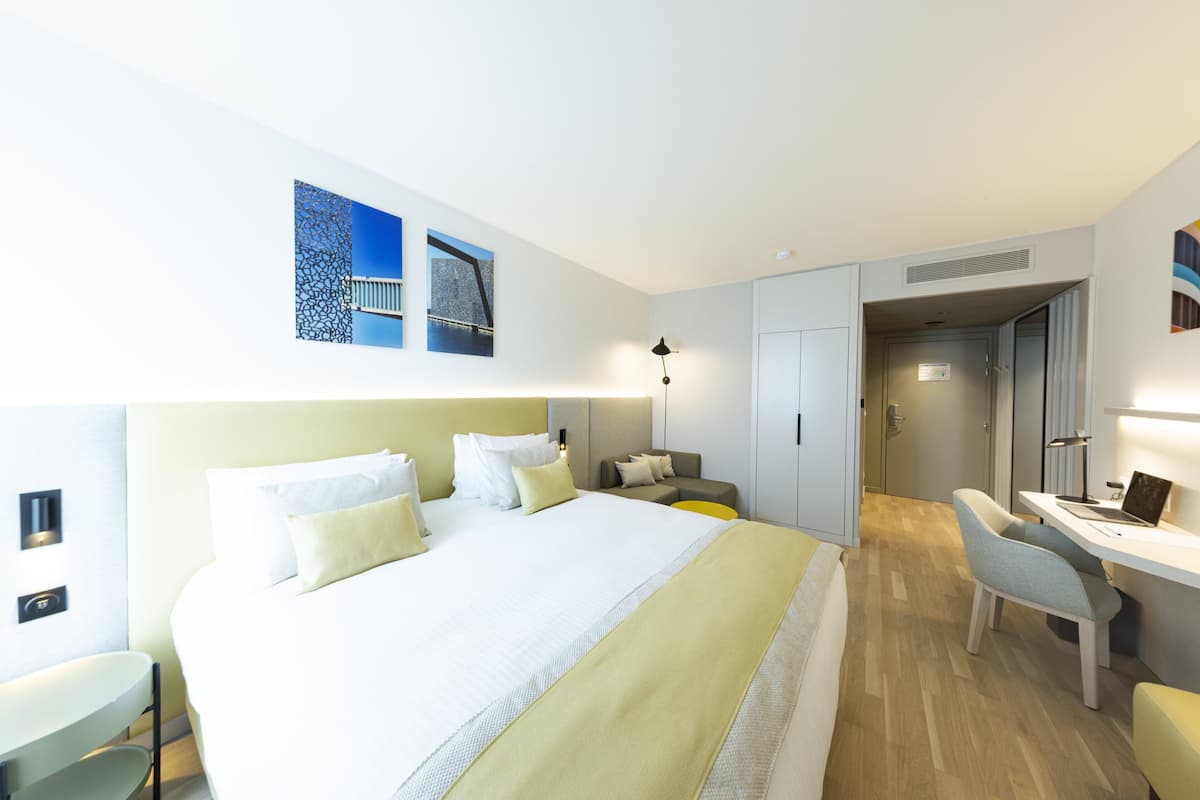 A modern hotel room with a large bed, a desk, and a chair. There is a door leading to another room and a small table with a lamp.