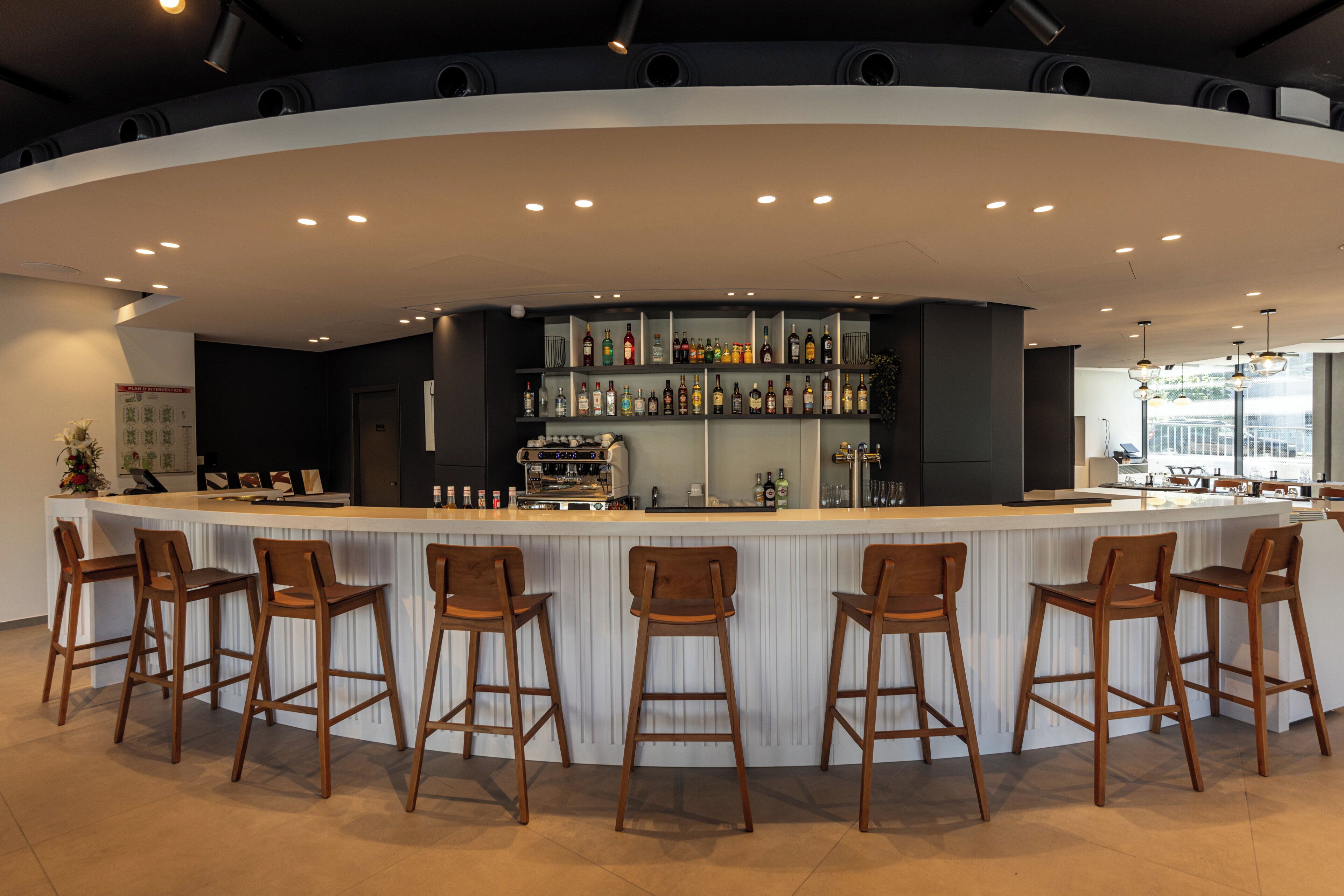 A modern bar area with a curved counter, multiple stools, and a variety of bottles and glasses on shelves.