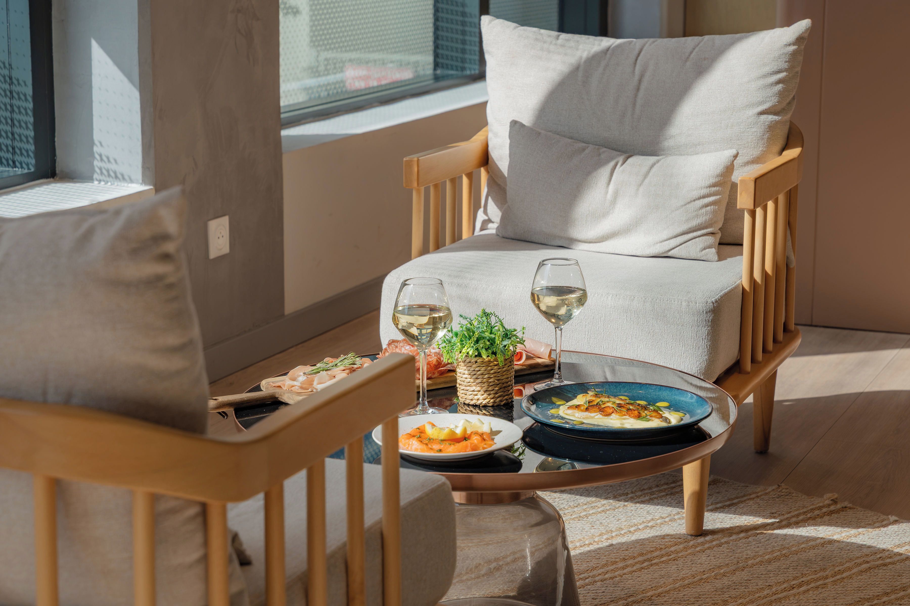 A modern living room with a wooden chair and sofa, a glass coffee table with food and drinks, and a view of the city through large windows.