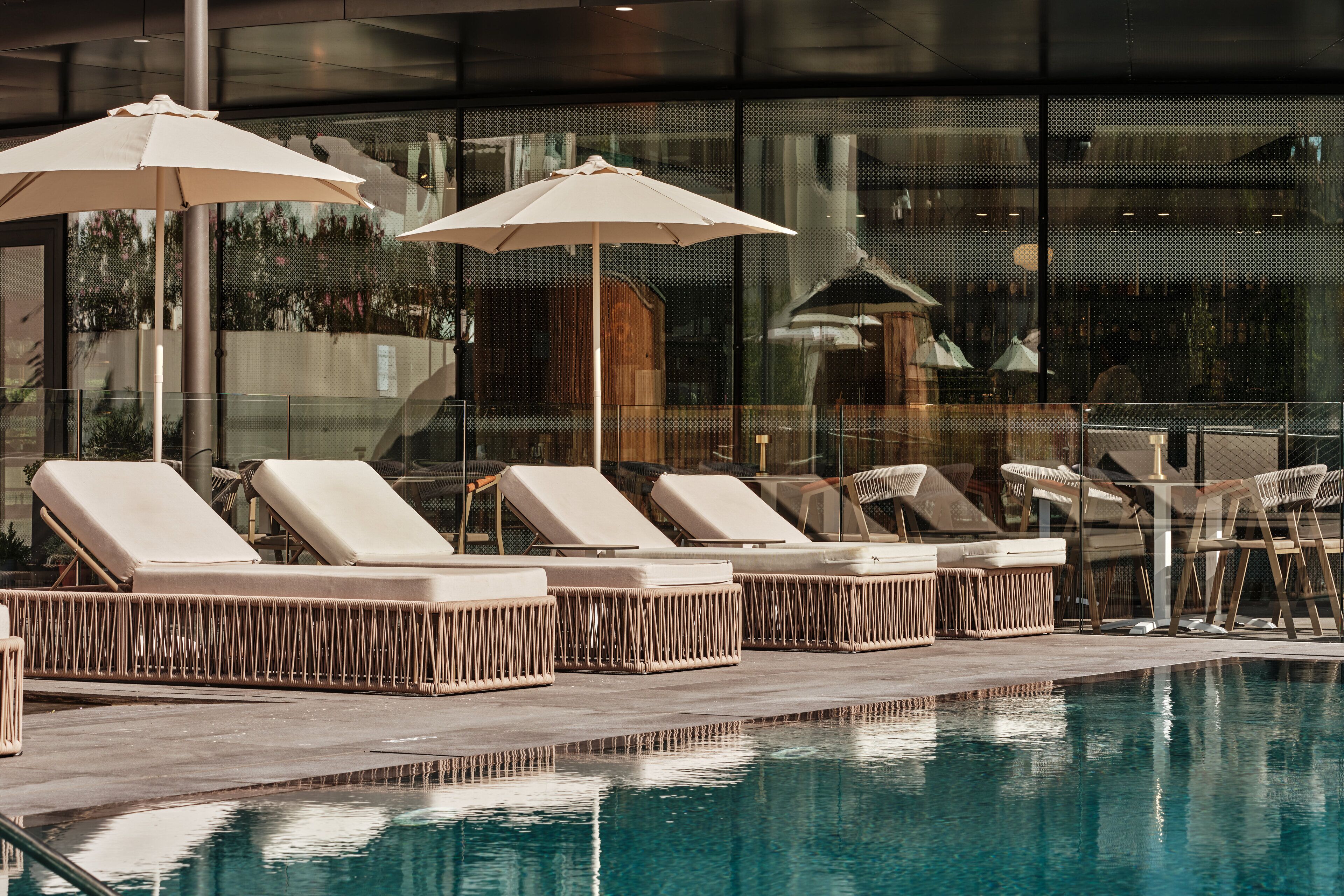 A poolside area with lounge chairs and umbrellas, a glass barrier, and a reflection of the building in the water.