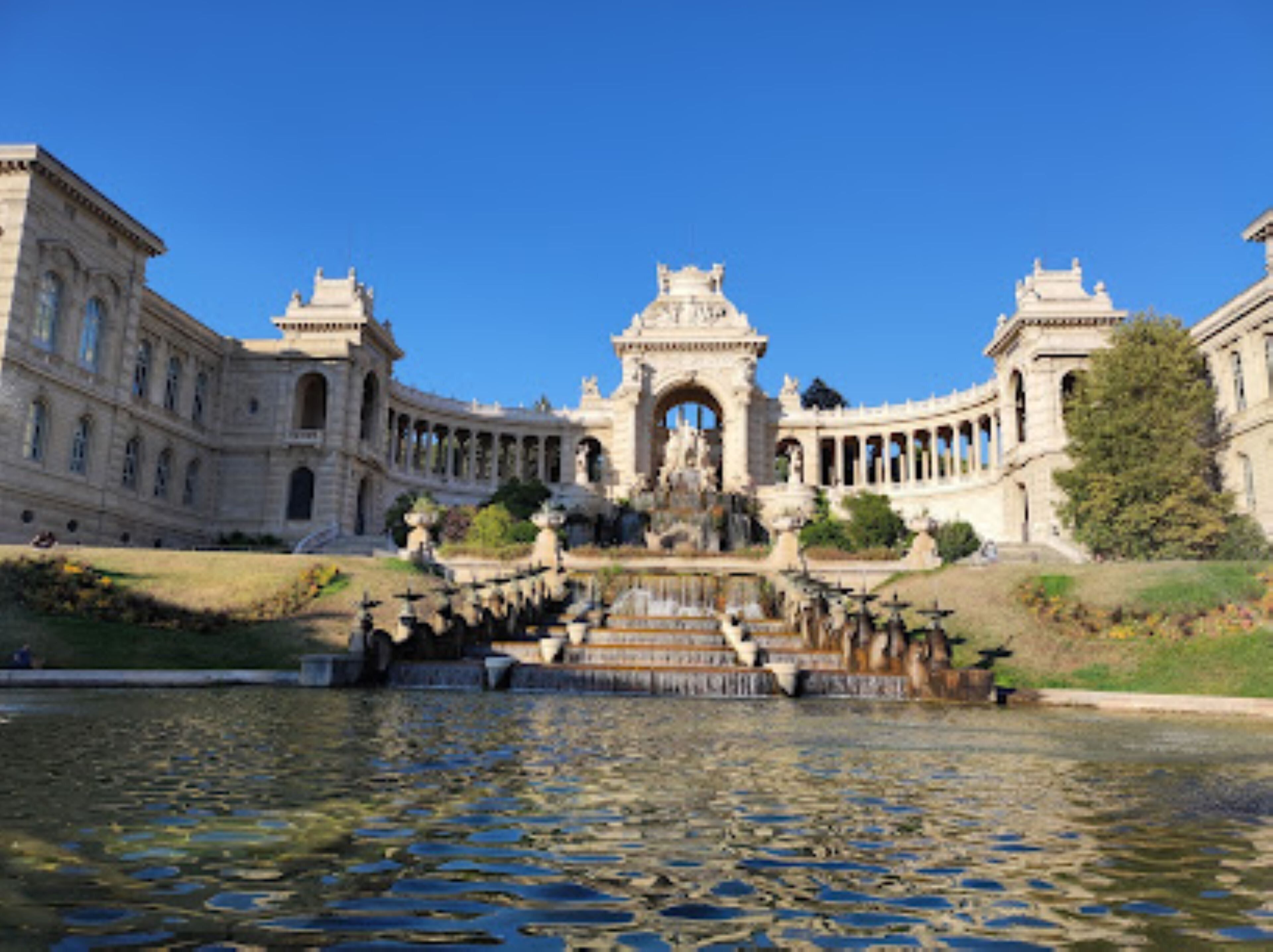 Un edificio imponente con un arco central, una gran fuente y un reflejo en el agua.