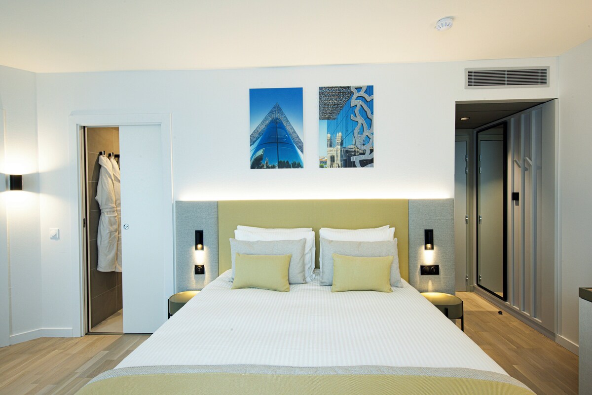 Habitación de hotel con una cama grande, dos mesitas de noche, un televisor montado en la pared y una puerta que da a un baño.