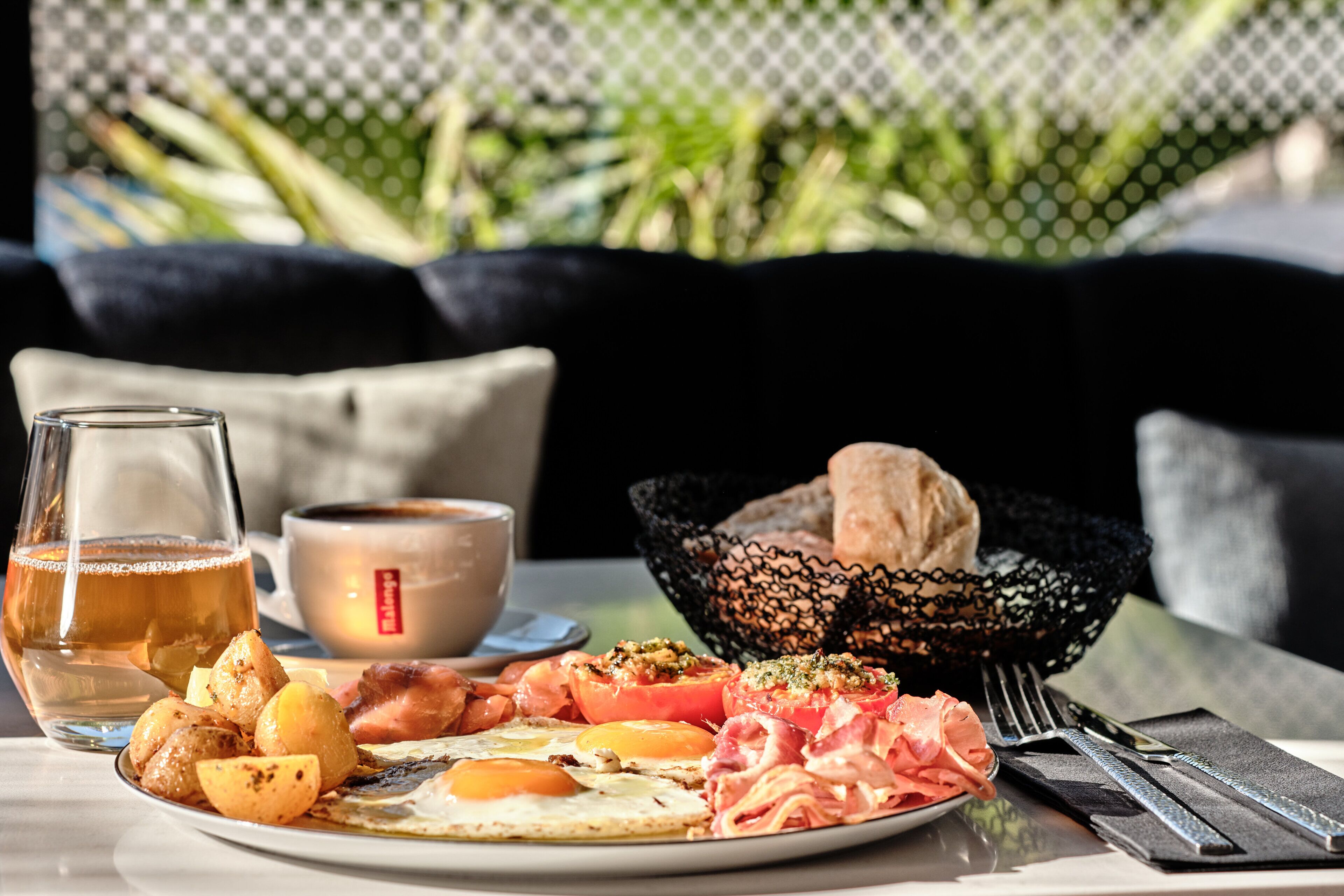 Un plat d'esmorzar amb ous, bacon, tomàquets i patates, un got de te i una tassa de cafè sobre una taula amb una cistella de fil negra i una tovallola.