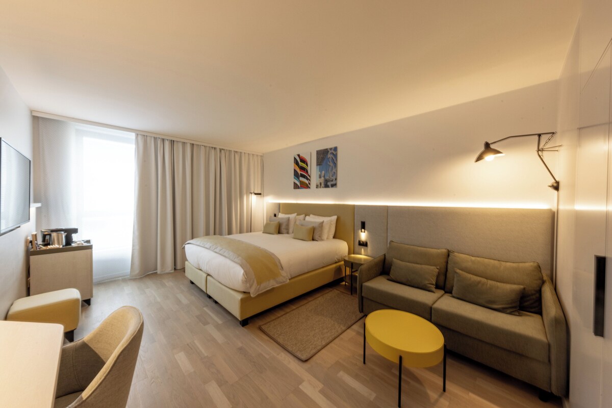 Una habitación de hotel moderna con una cama grande, un sofá, una mesita redonda y un escritorio con lámpara.