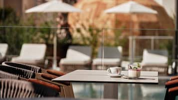 A poolside area with white lounge chairs and umbrellas, a table with a cup and a dessert, and a view of a building in the background.
