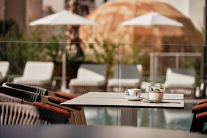 Zona junto a la piscina con sillones blancos y sombrillas, una mesa con una taza y un postre, y vista a un edificio en el fondo.