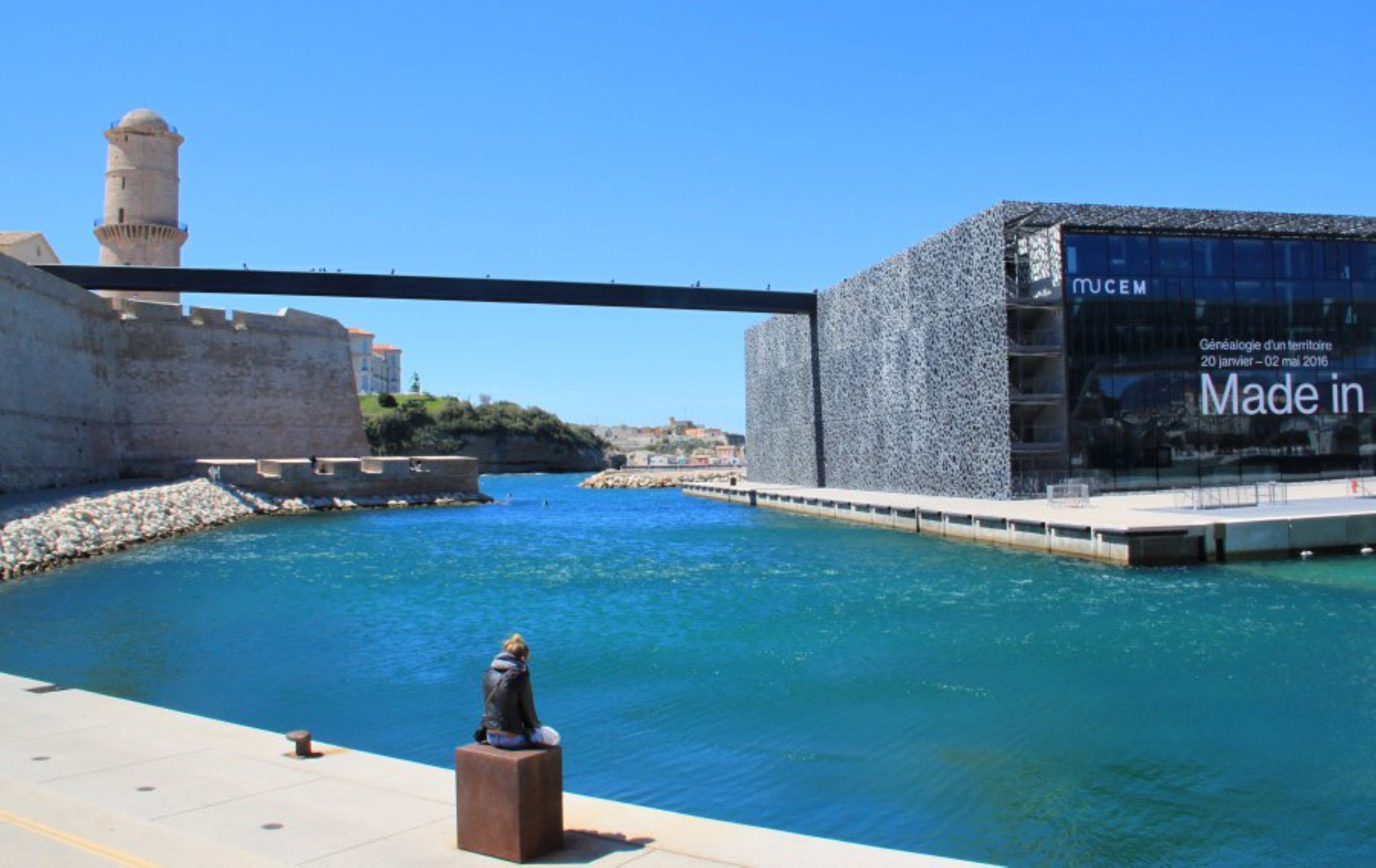Vista al agua con un edificio histórico, una estructura moderna con señalización 'Made in' y una estatua.
