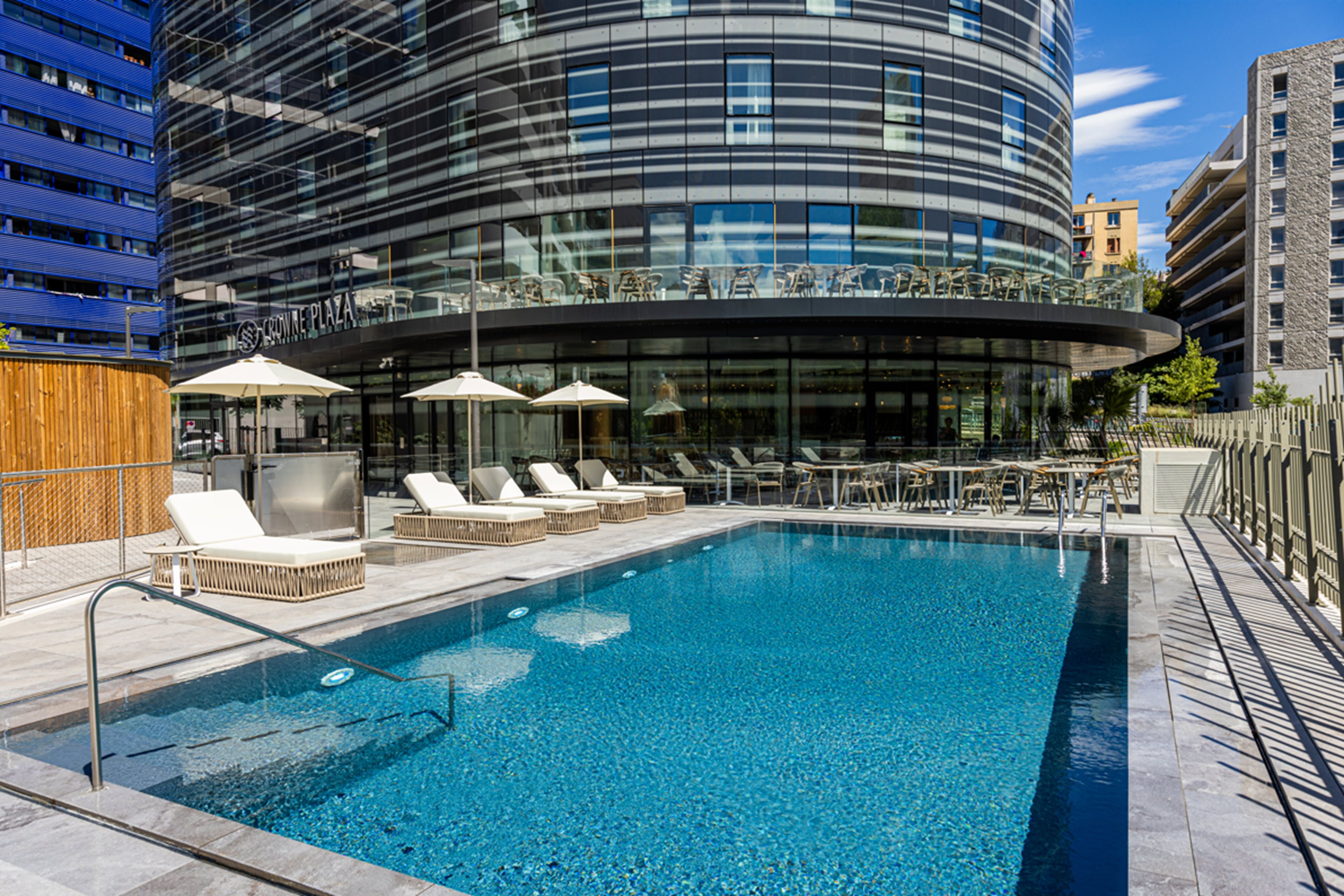 A modern building with a curved glass facade, a rooftop pool area with lounge chairs and umbrellas, and a clear blue sky.