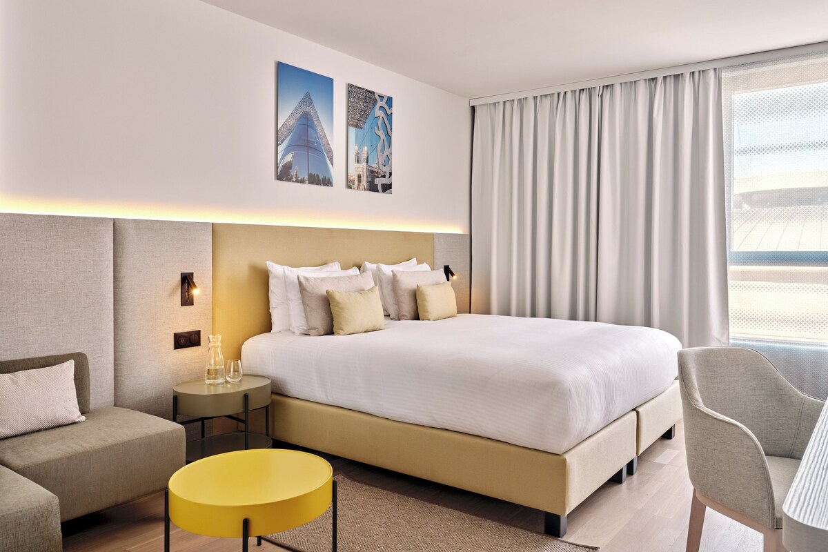 Habitación de hotel moderna con una cama grande, una mesita de noche con lámpara, una pequeña mesa redonda y una silla.