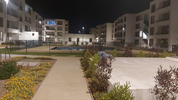 Vista nocturna de un complejo residencial con varios edificios de apartamentos, un sendero central y un área de juegos cercada.