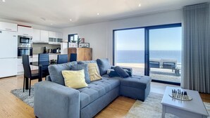 A modern living room with a grey sectional sofa, a dining area with a table and chairs, a kitchen with white cabinets, and a large sliding glass door leading to a balcony with a view of the sea. - Villa das Escaleiras (Porto Moniz)