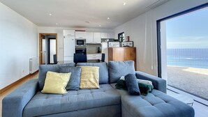A modern living room with a grey sectional sofa, a wooden cabinet, and a view of the sea through large windows. - Villa das Escaleiras (Porto Moniz)