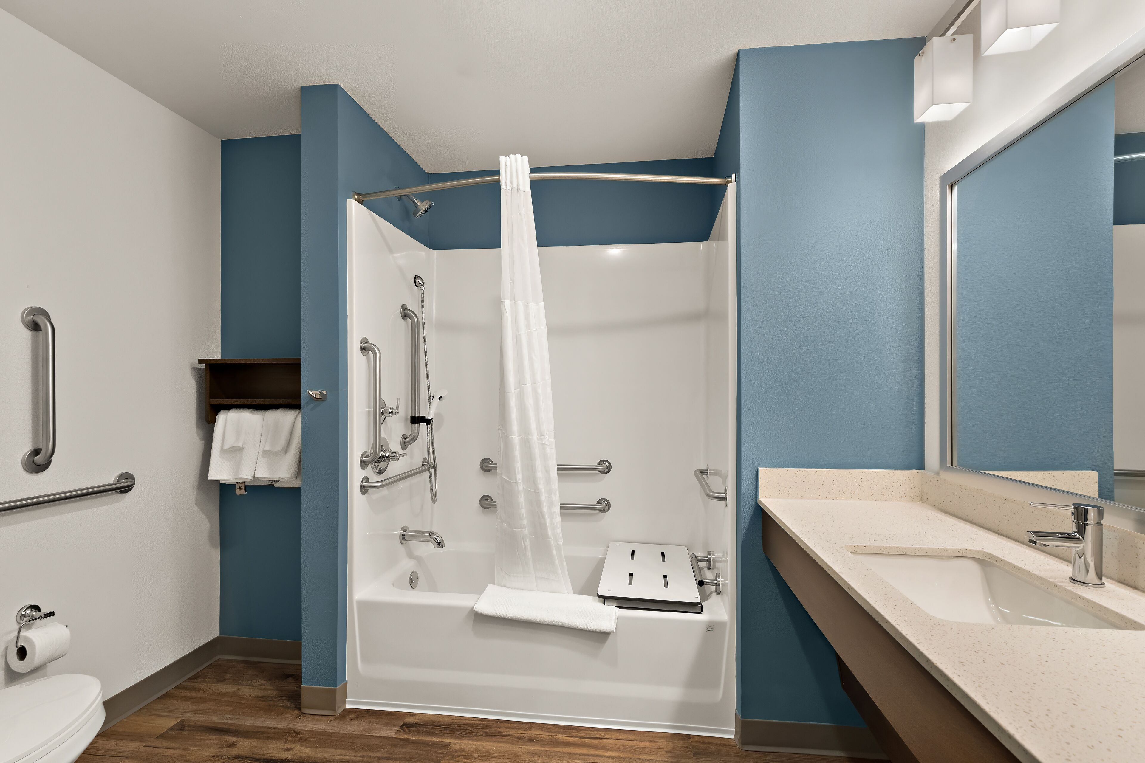 A modern bathroom with a bathtub, shower, and sink. It includes grab bars, a towel rack, and a mirror.