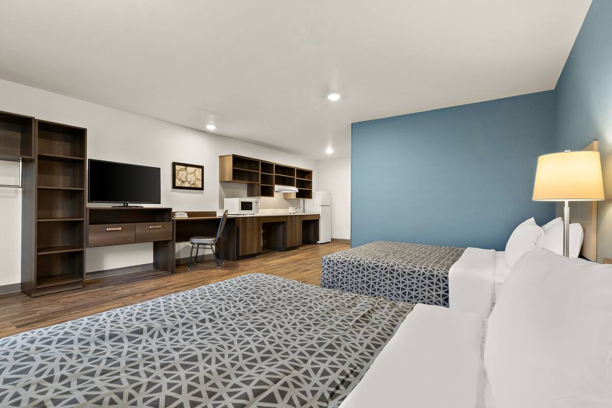 A modern hotel room with two beds, a desk, a TV, and a kitchenette.