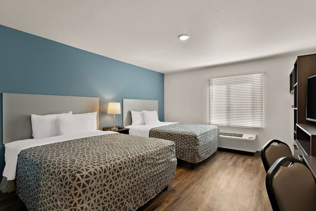 A hotel room with two beds, a TV, a desk, and a window with blinds.