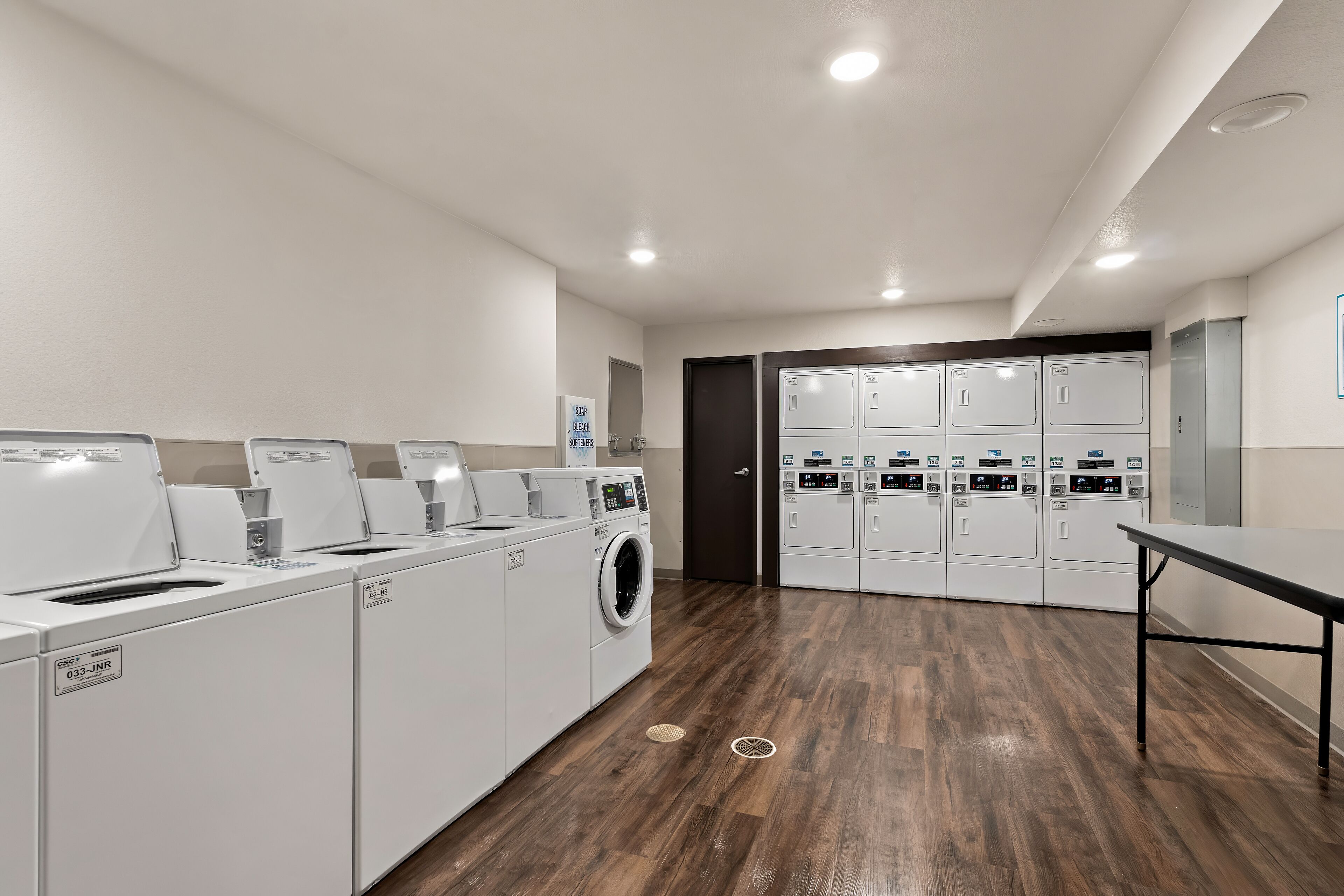 A laundry facility with multiple washing machines and dryers, a table, and a door leading to a storage area.