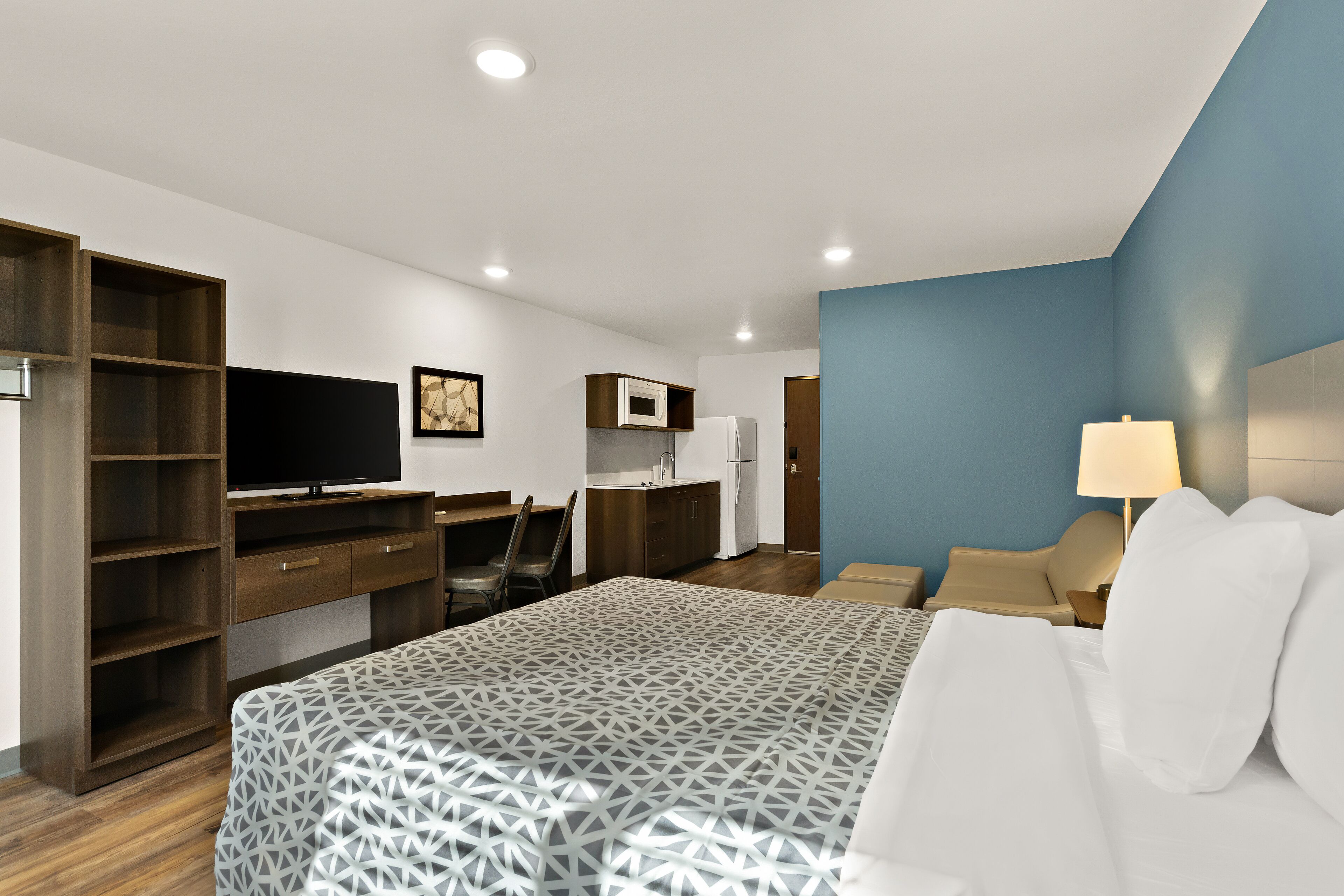 A hotel room with a bed, a desk, a television, a kitchenette, and a chair.