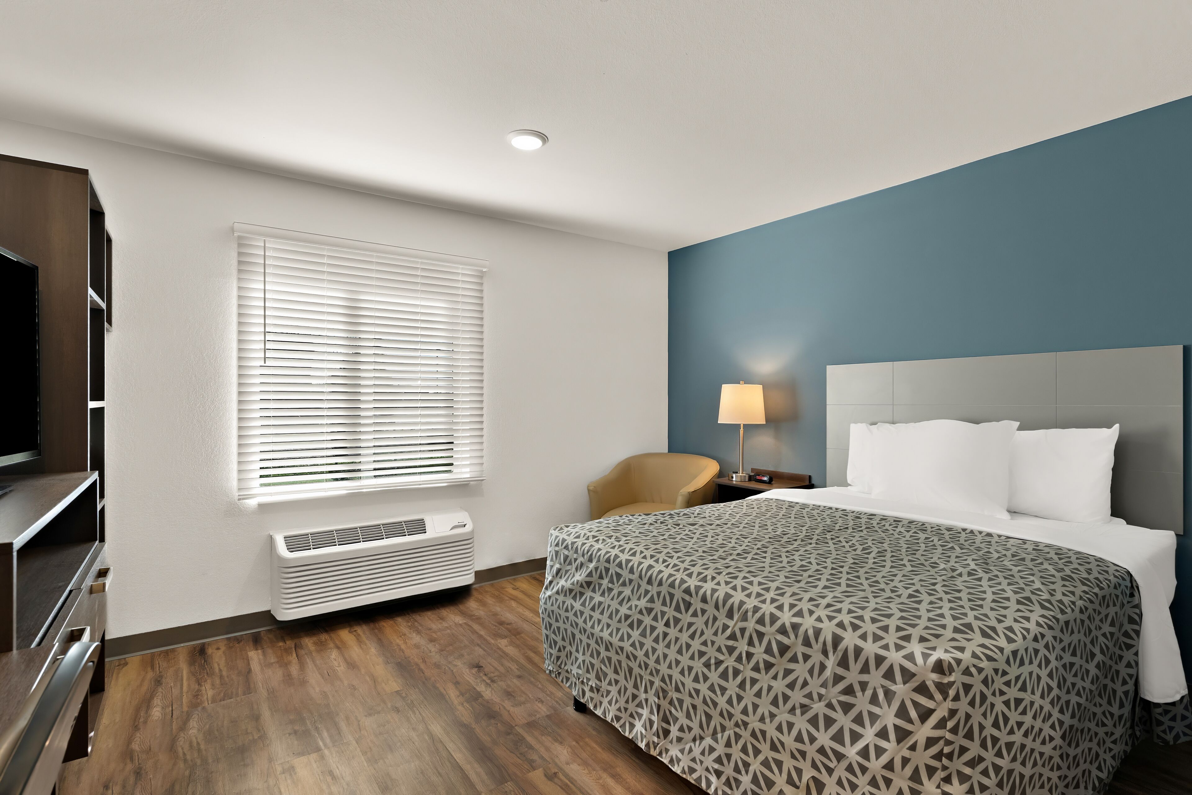 A hotel room with a bed, a TV, a desk, and a window with blinds.
