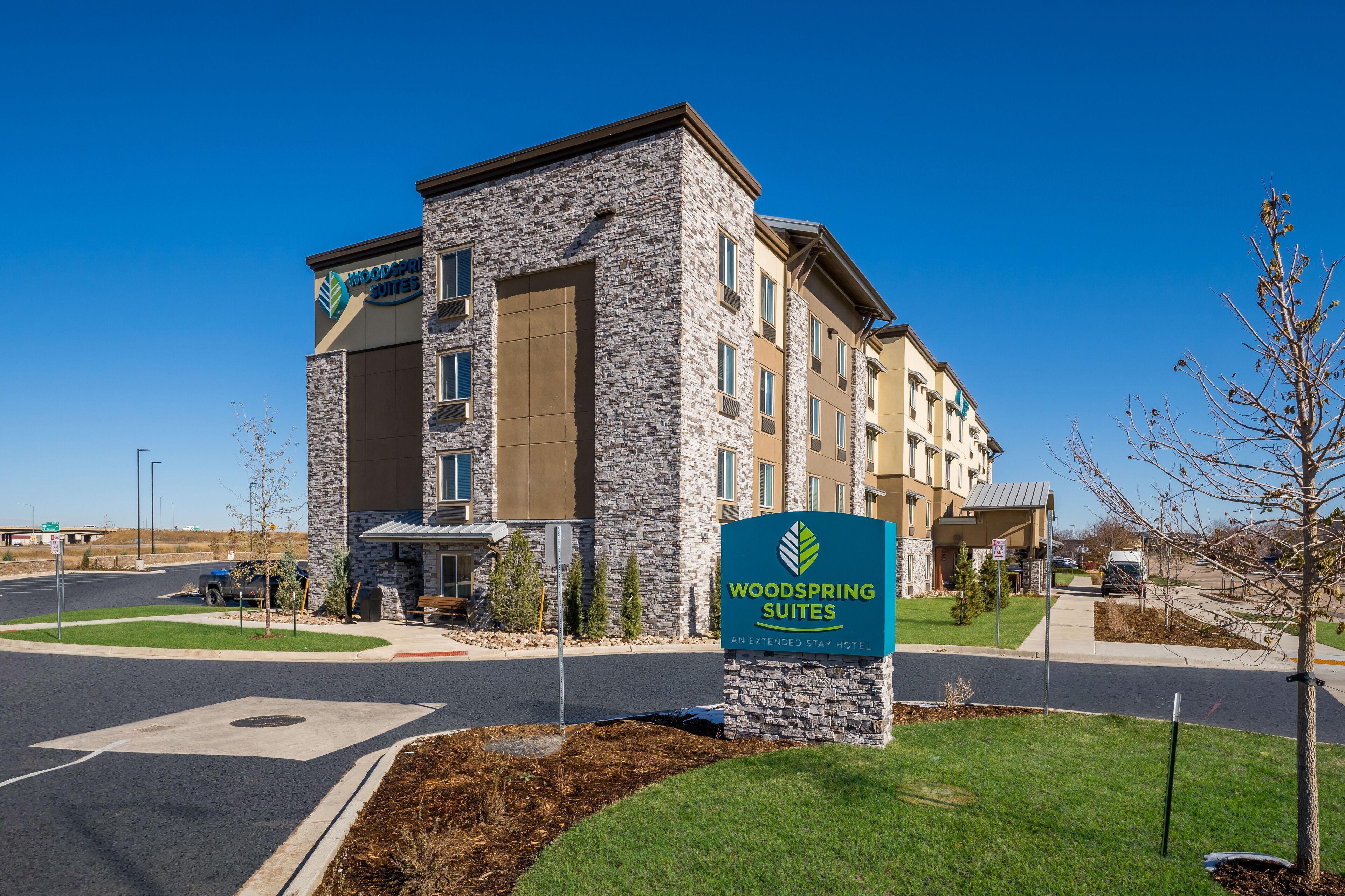 Un hotel de fachada de piedra, con un letrero que dice "Woodspring Suites" y un área ajardinada con acera y camino.