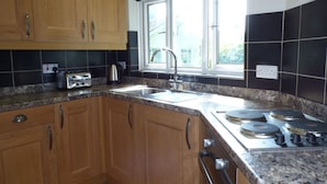 A kitchen with wooden cabinets, a granite countertop, a sink, a window, and a stove. - Spring Garden Cottage (Carmarthen)