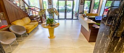 A hotel lobby with a wooden staircase, a reception desk, a yellow sofa, and a plant centerpiece.