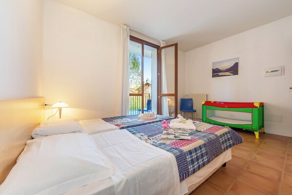 A hotel room with two beds, a window with curtains, a view of the outside, a children's playpen, and a painting on the wall.