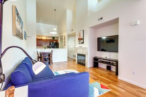 Uma sala de estar moderna com um sofá azul, piso de madeira, um espaço de jantar com cadeiras, uma cozinha ao fundo e uma TV na parede.
