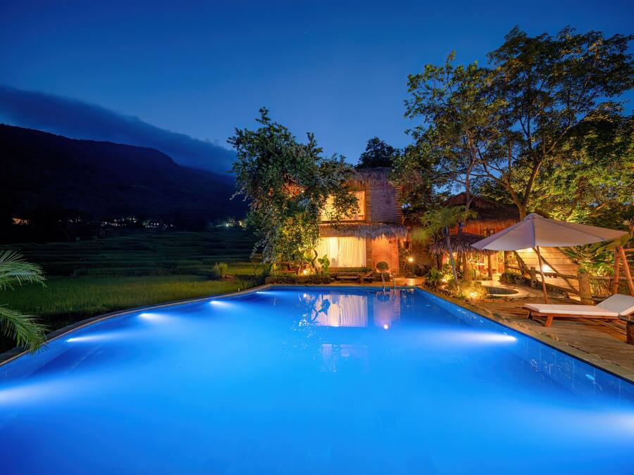 A swimming pool with lounge chairs and umbrellas, surrounded by a wooden deck and a thatched-roof building.