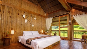 A wooden room with a large bed, a nightstand, a lamp, and a view of a green landscape through a glass door. - Puluong Luna Resort (Pu Luong)