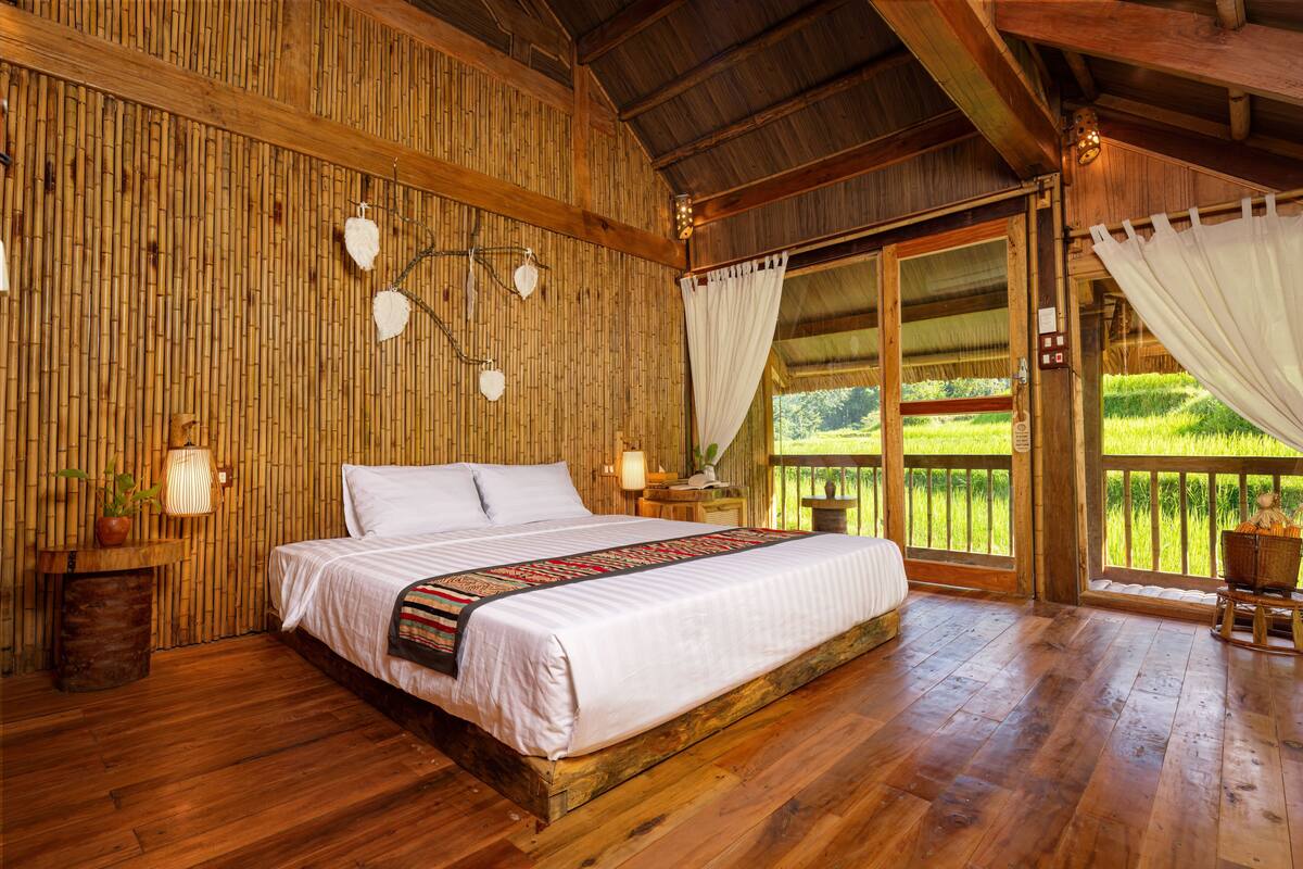 Ein Zimmer aus Holz mit einem großen Bett, einem Nachttisch, einer Lampe und Blick auf eine grüne Landschaft durch eine Glastür.