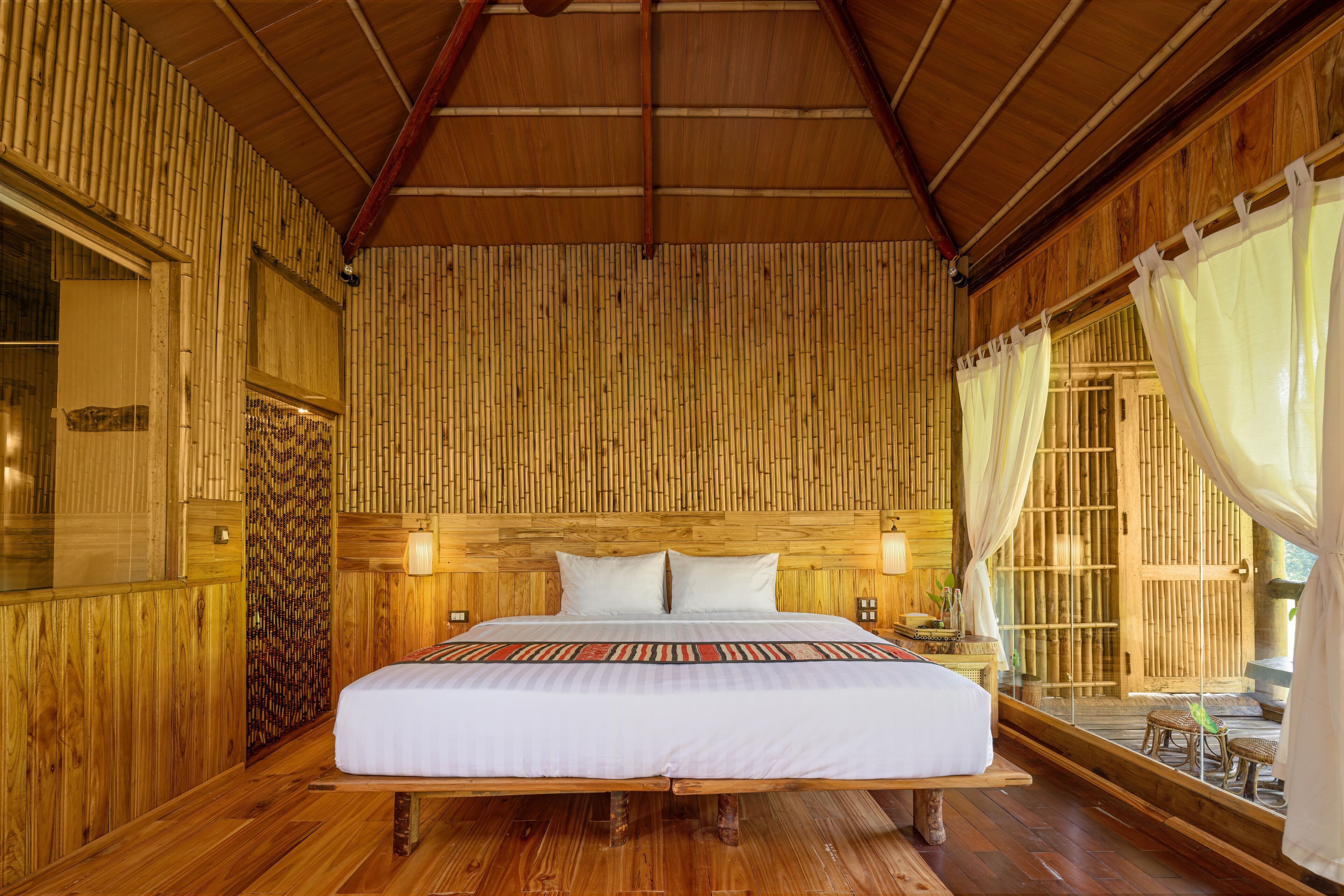 A large bed with a white bedspread and two pillows, wooden walls, a wooden ceiling, and a window with white curtains.