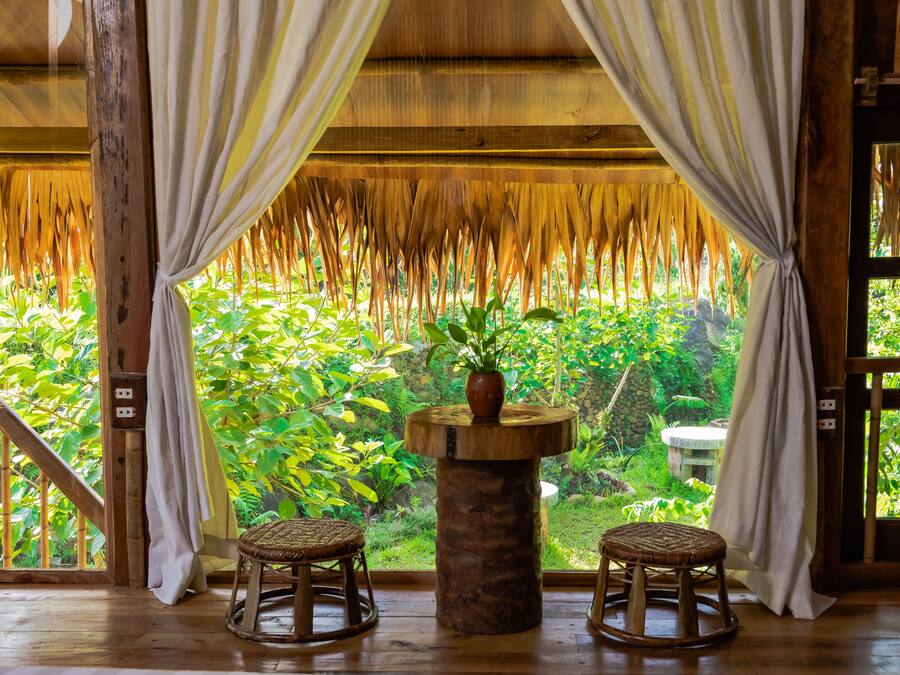 A room with a wooden ceiling, a table with a plant, two wicker stools, and a view of lush greenery through large windows.