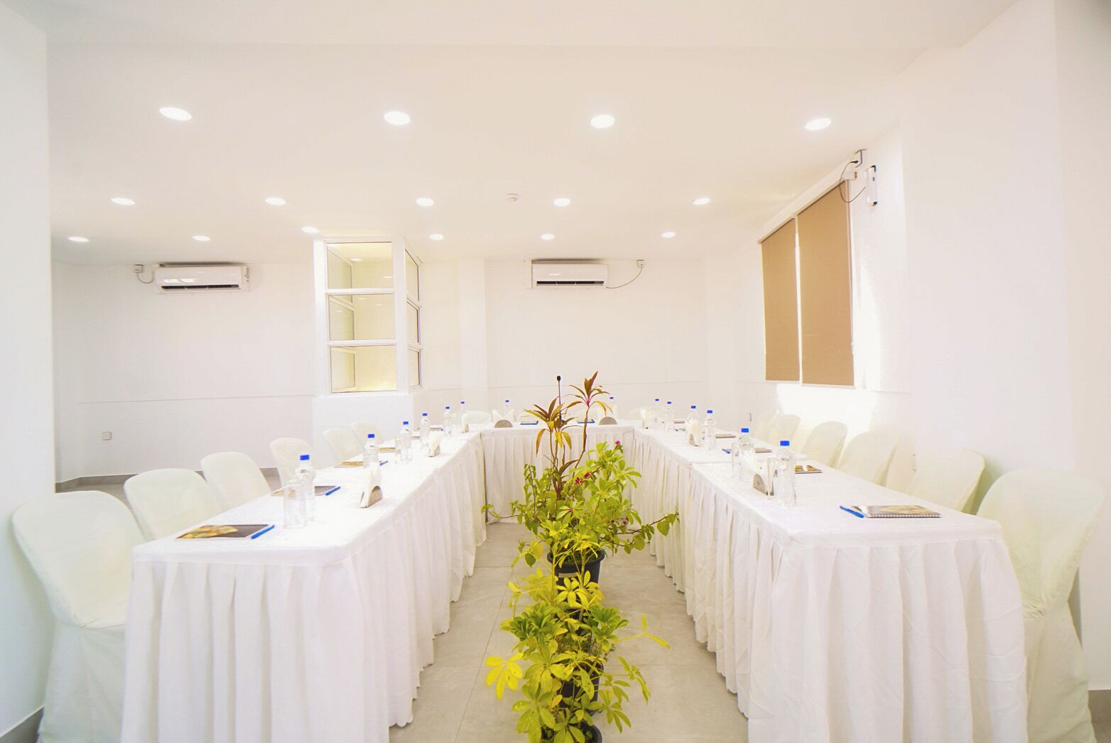A conference room with white tables and chairs, a central plant, and air conditioning units on the walls.