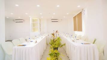 A conference room with white tables and chairs, a central plant, and air conditioning units on the walls.