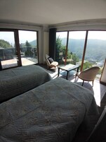 A bedroom with two beds, a chair, a small table, and large windows offering a mountain view.