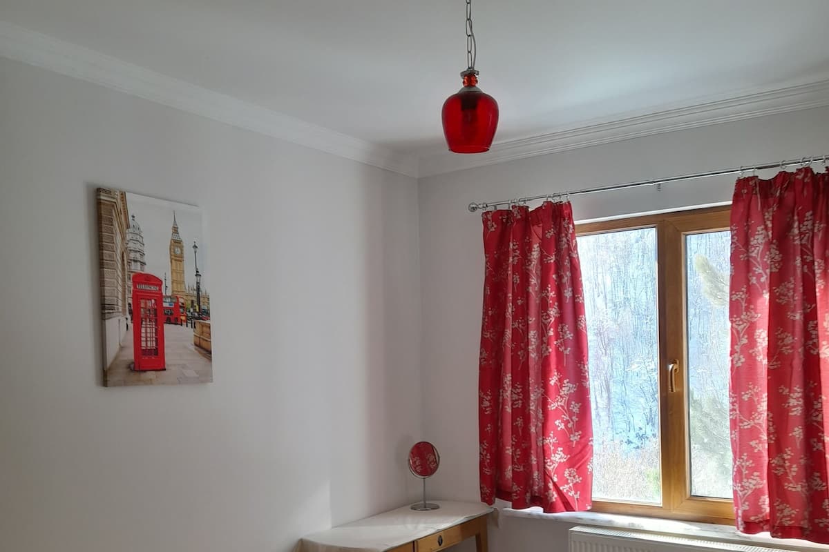 A bedroom with a bed, a bedside table, a window with red curtains, a radiator, and a wall-mounted picture of a London landmark.