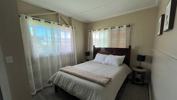 A hotel room with a bed, two bedside tables with lamps, a window with sheer curtains, and a view of greenery outside.