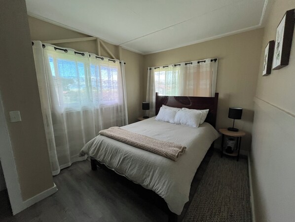 A hotel room with a bed, two bedside tables with lamps, a window with sheer curtains, and a view of greenery outside.