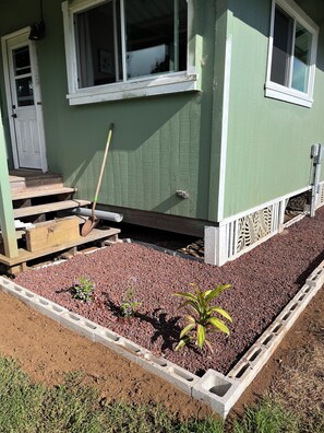 A house with a green exterior, a wooden porch, and a garden bed with red soil and plants.