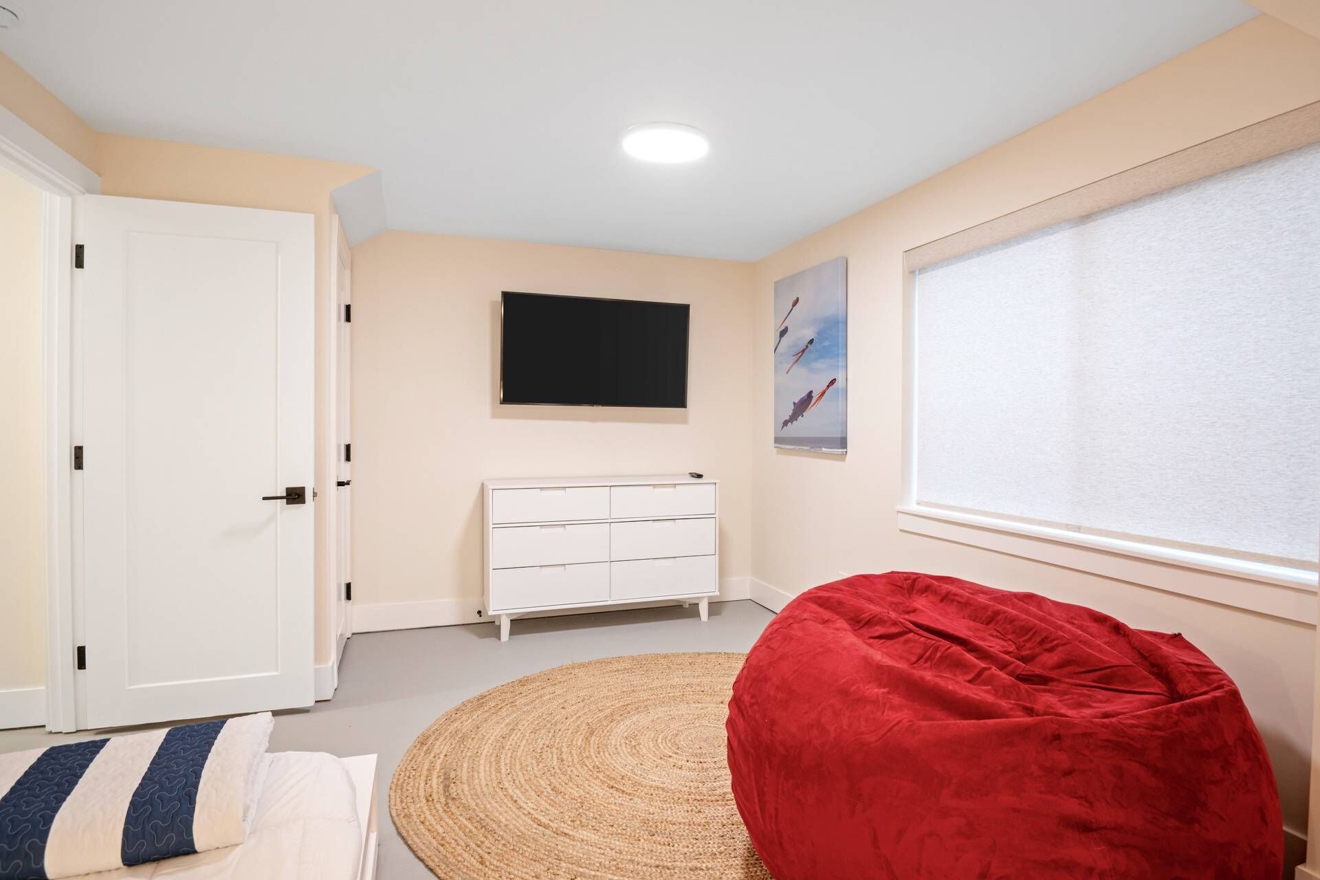 Un dormitorio con cama, un sillón de mimbre rojo, un aparador blanco, un televisor de pantalla plana, una ventana con persianas y una obra de arte colgada en la pared.