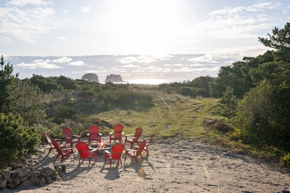 A sandy beach with red chairs arranged around a central fire pit, surrounded by greenery and distant rock formations. - Magnificent Oceanfront Views of Twin Rocks on Private Acre, Pet Friendly, Hot Tub, Fire-Pit, Deck (Rockaway Beach)