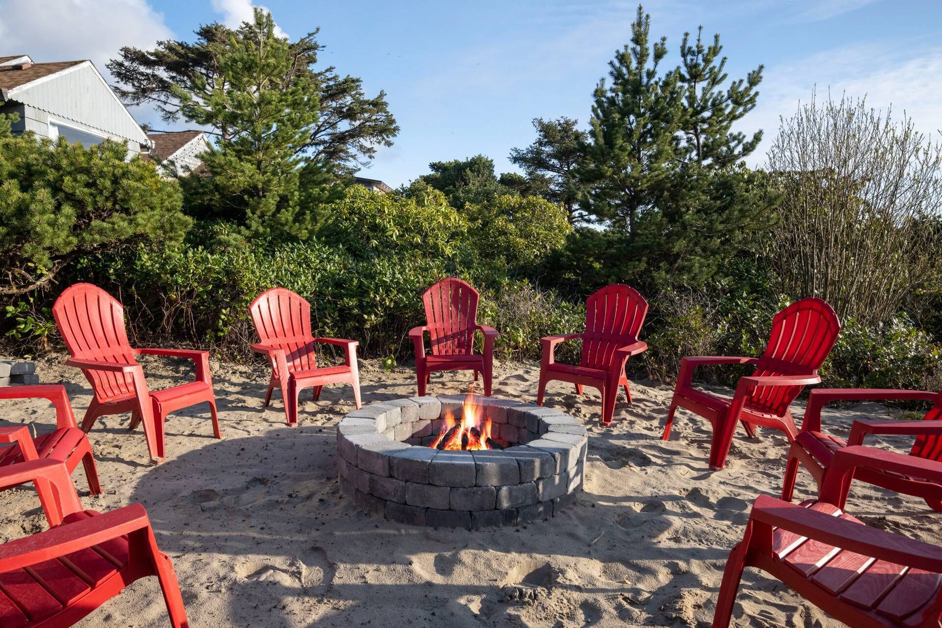 A circular fire pit surrounded by red Adirondack chairs on a sandy area with trees and a building in the background.