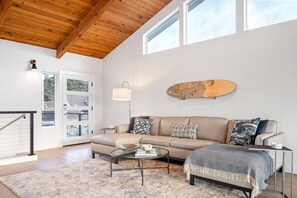 A modern living room with a wooden ceiling, a beige sofa, a glass coffee table, and a skateboard wall art piece.