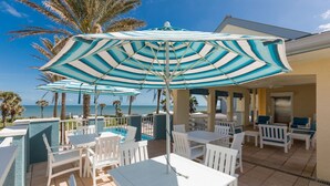 A poolside area with white chairs and tables, a large striped umbrella, and a view of the ocean. - SouthernVilla Ticket to Paradise (Palm Coast)