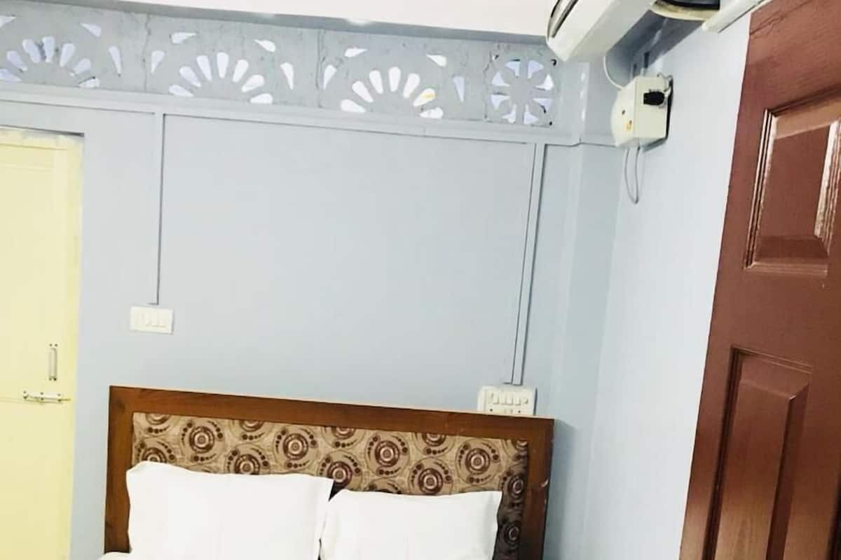 A neatly made bed with white sheets and a beige blanket, a wooden headboard with a patterned design, and a wall-mounted air conditioning unit.