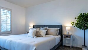 A bedroom with a large bed, two bedside tables with lamps, a potted plant, and a window with blinds. - Lux Haven with Movie Theater, Hot Tub, Office, and Grill NestNights (Santa Clarita)