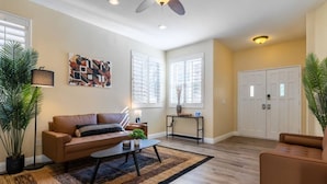 A living room with a brown leather sofa, a coffee table, a floor lamp, and a painting on the wall. - Lux Haven with Movie Theater, Hot Tub, Office, and Grill NestNights (Santa Clarita)