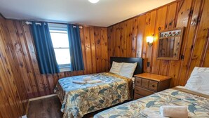 Una habitación con paneles de madera, dos camas con cubrecamas florales, una mesita de noche y un aplique de pared.